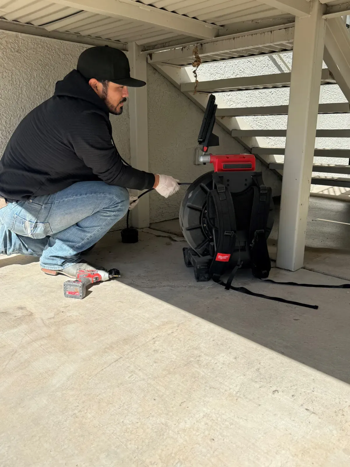 Tucson Plumbing technician feeding a sewer camera from a backpack reel into a cleanout beneath an outdoor staircase